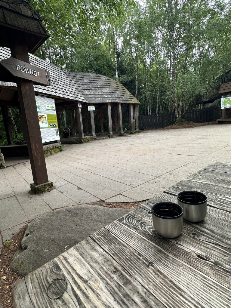 Stół i na nim dwa kubki z kawą. W tle kryte zadaszenie na końcu ścieżki dydaktycznej